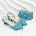 A pair of sparkling turquoise blue pointed-toe slingback flats with layered flower embellishments on the toes, shown alongside a matching glittery clutch purse with a gold-tone chain clasp on a white wavy background, Obroi