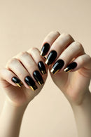 A close-up of hands with glossy black coffin-shaped nails featuring a metallic gold French tip design, set against a soft beige background, Obroi