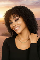Woman with curly hair wearing a black top and gold necklace against a light gray background, Obroi