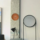 A modern wall clock with a terracotta orange face and black hands, mounted on a textured gray concrete pillar in a minimalist interior setting next to a black-framed floor mirror, Obroi
