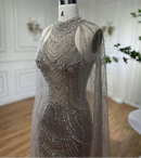 A sleeveless, high-neck evening gown covered in intricate silver beadwork and sequins, displayed on a mannequin. The dress features a sheer, matching mesh cape draped over the shoulders and arms, Obroi