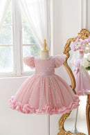 A pink toddler girl's party dress displayed on a mannequin dress form, featuring puff sleeves, a pearl-embellished bodice, and a ruffled tulle skirt, with a gold mirror reflecting the back of the dress in a bright room, Obroi