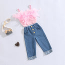 A children's outfit set laid flat on a light background, featuring a pink feather-trimmed camisole top, blue denim capri pants with three front buttons, a small beige camera prop, and a small heart-shaped ornament, Obroi