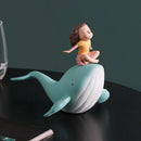 A decorative figurine of a child sitting on a whale, placed on a dark tabletop with a dark green background. The child wears a yellow top and pink skirt, and the whale is light blue and white, Obroi