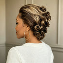 A woman shown from the side and back wearing a styled updo wig with defined curls and highlighted blonde streaks. She is wearing a white knit sweater and large gold hoop earrings, photographed indoors against a neutral paneled wall, Obroi