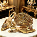 A luxury gold bridal shoe set featuring pointed-toe slingback heels with crystal embellishments, a matching round beaded clutch purse, and a matching gold belt bag, displayed on a white marble table in an ornate, chandelier-filled room, Obroi