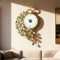 A decorative wall clock shaped like a peacock, with a round white face, gold-tone frame and numerals, and cascading peacock-feather details in gold, clear beads, and turquoise-blue stones. The clock is mounted on a beige wall in a sunlit room, with a sofa and framed artwork partially visible below and to the right, Obroi