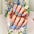 A close-up of a hand with light blue and floral nail art, resting on a matching floral-patterned surface with lace trim. The background is softly blurred with decorative flowers and a chandelier, Obroi