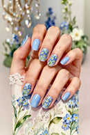 A close-up of a hand with light blue and floral nail art, resting on a matching floral-patterned surface with lace trim. The background is softly blurred with decorative flowers and a chandelier, Obroi