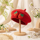 Red knitted beret with strawberry decorations on a wooden stand, surrounded by flowers and plants, Obroi