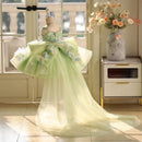 A formal girl's party dress displayed on a mannequin in a bright room. The dress is pale mint green with a beaded bodice, a large ruffled bow at the waist, and floral appliqués in light blue and white, Obroi