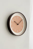 A minimalist round wall clock with a dark brown frame, a warm peach-pink dial, and white numerals, mounted on a light gray wall, Obroi