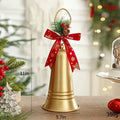 A gold bell-shaped Christmas ornament with a red ribbon bow and green pine sprigs, shown on a tabletop with holiday decorations in a softly blurred background. Measurement labels reading 28cm, 14, Obroi