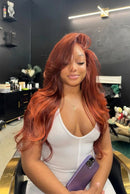 A woman with long, wavy coppery red hair sits in a salon chair wearing a white top and holding a purple phone. The background shows a dark salon interior with shelves and fixtures, Obroi