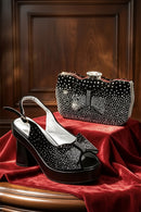 A black platform slingback high heel shoe with a rhinestone bow detail, displayed next to a matching rhinestone-embellished clutch bag on a red velvet surface against a dark wood background, Obroi