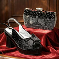 A black platform slingback pump with a rhinestone bow detail and a matching black clutch bag with a rhinestone bow, displayed on a red velvet surface against a dark wood background, Obroi