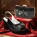 A black platform slingback pump with a rhinestone bow detail and a matching black clutch bag with a rhinestone bow, displayed on a red velvet surface against a dark wood background, Obroi