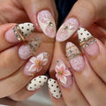 Close-up of manicured hands showcasing a variety of elegant nail art designs. The nails feature white and pink designs including lace patterns, 3D flowers, polka dots, rhinestones, and gold rings, Obroi
