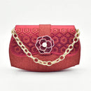 A red textured clutch handbag with a gold chain strap, featuring a central rose-shaped embellishment with a pearl center and geometric patterns on the flap, Obroi