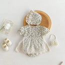A baby girl's short-sleeve floral romper dress with a matching bonnet, styled in a flat lay with baby shoes, a pearl purse, and a flower on a light background, Obroi