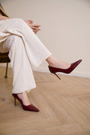 A person seated on a chair wearing glossy deep burgundy pointed-toe high heels with slim stiletto heels, paired with cream wide-leg trousers, in a room with light wood flooring and a beige wall, Obroi