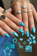 A close-up of manicured fingernails with a glossy metallic turquoise-blue leaf pattern, shown on a person's hands wearing multiple rings. Decorative nail art sample graphics appear at the bottom, Obroi