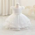 A white baby girl's formal dress displayed on a mannequin, featuring a textured lace bodice, short cap sleeves, a matching waist bow, and a tiered tulle skirt with lace trim, Obroi