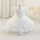 A white baby girl's formal dress displayed on a mannequin, featuring a textured lace bodice, short cap sleeves, a matching waist bow, and a tiered tulle skirt with lace trim, Obroi