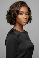 A woman with a short, styled brown wig with soft curls, shown in a three-quarter portrait against a solid gray background, Obroi