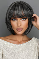 A woman wearing a short, layered wig with straight-cut bangs. The wig has a dark charcoal gray base with lighter ash gray highlights, styled against a solid gray background, Obroi