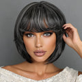 A woman wearing a short, layered wig with straight-cut bangs. The wig has a dark charcoal gray base with lighter ash gray highlights, styled against a solid gray background, Obroi