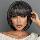 A woman wearing a short, layered wig with straight-cut bangs. The wig has a dark charcoal gray base with lighter ash gray highlights, styled against a solid gray background, Obroi