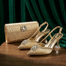 A pair of gold pointed-toe slingback heels with crystal embellishments and a matching gold clutch bag, displayed on a wooden table against a dark green velvet background, Obroi