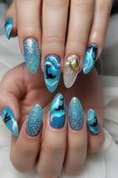 A close-up of hands showcasing a detailed ocean-themed nail art design. The nails feature various designs including blue and white waves, black dolphin silhouettes, glittery blue sections, and two accent nails decorated with gold seashells and white faux pearls, Obroi