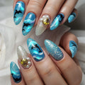 A close-up of hands showcasing a detailed ocean-themed nail art design. The nails feature various designs including blue and white waves, black dolphins, glittery blue sections, and two silver glitter accent nails decorated with gold seashells and white faux pearls, Obroi