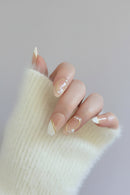 A close-up of a hand wearing a fluffy cream knit sleeve and elegant almond-shaped nails with white and gold French tip designs and pearl accents, set against a soft gray background with a magazine page in the back, Obroi