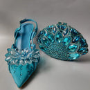 Blue high-heeled shoe with crystal embellishments and a matching blue handbag on a gray background, Obroi