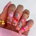 Hand with pink nail art featuring hearts, flowers, and text on a white background, Obroi