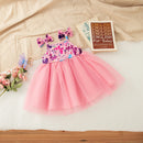A toddler girl's sleeveless tulle dress with a floral patterned bodice and a voluminous pink tulle skirt, styled in a flat-lay arrangement with a matching bow headband, flowers, and a book on a beige fabric background, Obroi