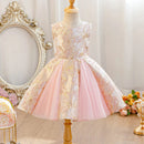 A sleeveless children's party dress displayed on a dress form. The dress has a champagne-gold and white floral brocade bodice with a matching waistband, and a flared light pink tulle skirt, Obroi