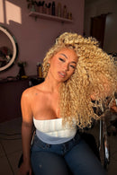 A woman with voluminous, tightly curled blonde hair wearing a white strapless top, posing in a salon setting with a mirror and salon decor visible in the background, Obroi