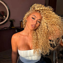 A woman with voluminous, tightly curled blonde hair wearing a white strapless top, posing in a salon setting with a mirror and styling tools visible in the background, Obroi