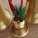 A close-up of a gold bell-shaped holiday ornament or candle holder with a red ribbon, green pine needles, and red berries on a neutral surface, Obroi