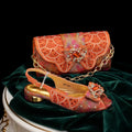 An orange embellished evening bag and matching heeled sandal displayed together on dark green velvet fabric against a black background. The accessories feature intricate beadwork, rhinestone patterns, gold-tone chain details, and a jeweled orange accent on the flap and sandal strap, Obroi