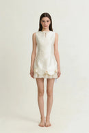 A woman stands barefoot against a plain light background wearing a sleeveless cream-colored mini dress with a bow at the neckline and decorative ruffle details near the hem, Obroi