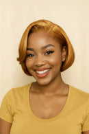 A smiling woman with a short, wavy golden-blonde bob wig, wearing a mustard yellow scoop-neck t-shirt and a delicate gold necklace, set against a plain cream background, Obroi