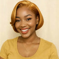 A smiling woman with a short, wavy golden-blonde bob wig, wearing a mustard yellow scoop-neck t-shirt and a delicate gold necklace, set against a plain cream background, Obroi