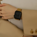 A close-up of a person's wrist wearing a smartwatch with a black rectangular face and a shiny gold mesh band, resting against a beige blazer and a white bag with black lettering, Obroi