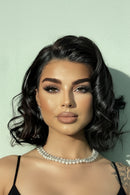A close-up portrait of a woman with styled dark wavy hair, dramatic eye makeup, and a pearl necklace against a pale mint wall, Obroi