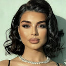 A close-up portrait of a woman with styled dark wavy hair, dramatic eye makeup, and a pearl necklace against a pale mint wall, Obroi